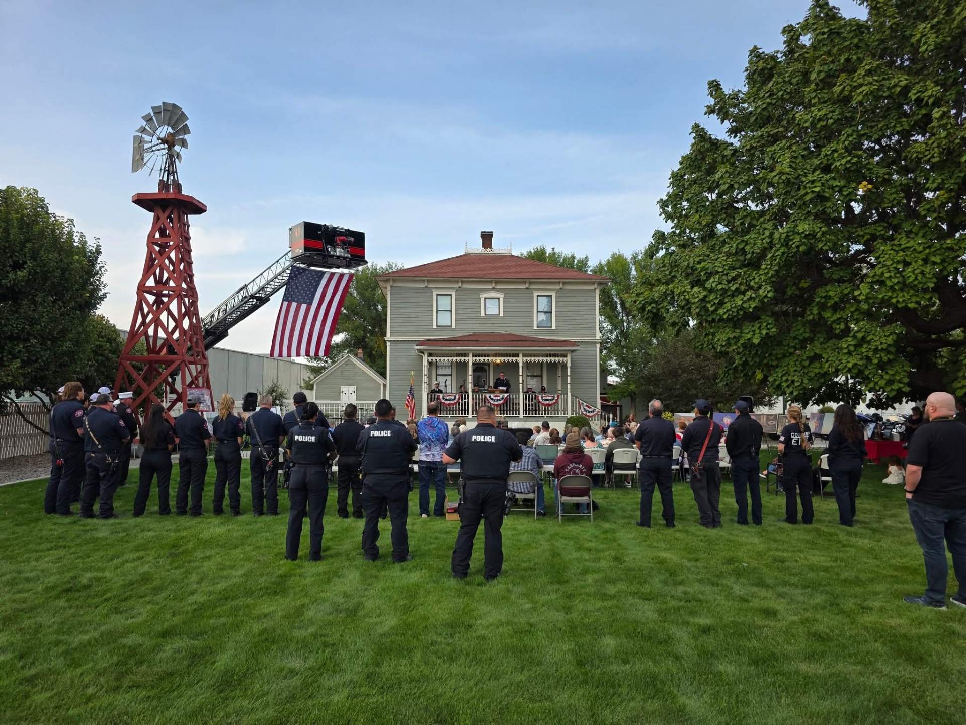 Police officers, firefighters and neighbors gathered at a memorial ceremony beside a historic house, windmill and ladder-truck flag